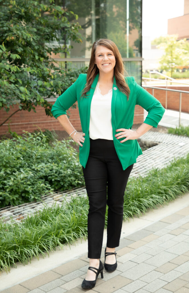 Amanda Knapp Full Body Image, wearing a green blazer in front of a building with green plants